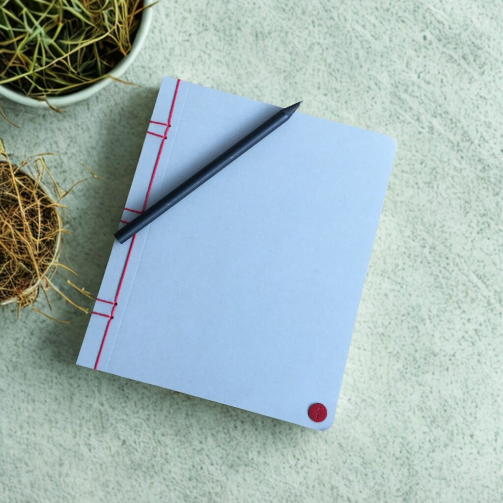Carnet Les Carnets de Lucie gris perle posé sur un bureau avec un crayon, reliure japonaise cousue main, papier 100 g/m² FSC.